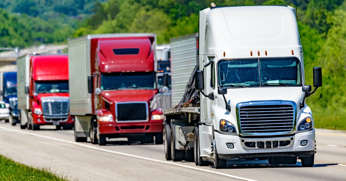 A white 18-wheeler truck leads a line of different colored semitrailers down a highway with trees on the side of the road.