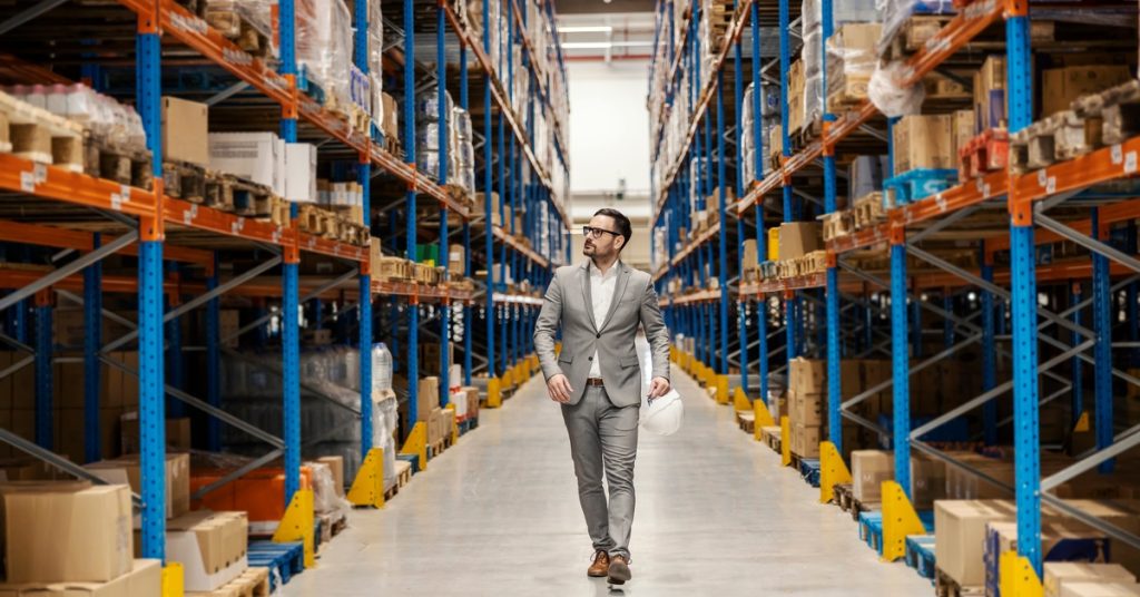 A man in a gray suit walks down an aisle in a warehouse with shelving full of cardboard boxes of products on both sides.