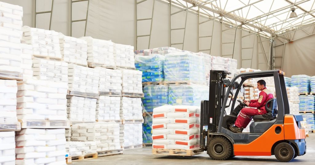 A worker operates an orange pallet fork loader machine with products stacked on it inside a warehouse.