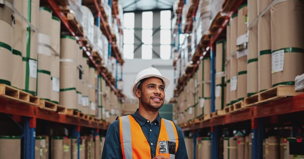 A smiling man wears a safety vest and hard hat while working in a warehouse filled with products on pallet racks.