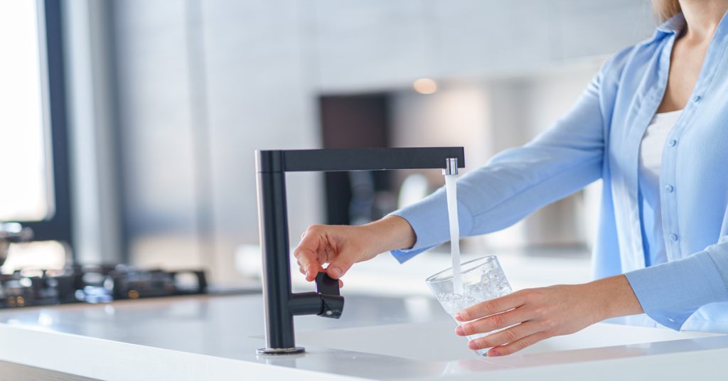 A woman in a blue button-down shirt fills a drinking glass with clean water from a black kitchen faucet.
