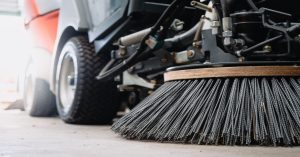 The round black bristle brush of a street sweeping vehicle rests on the ground in front of the wheels.
