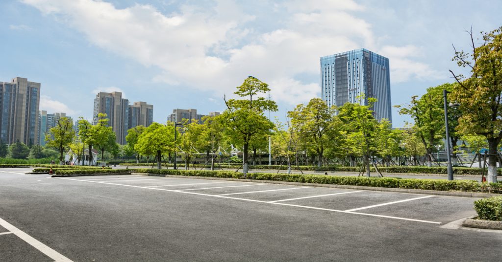 An empty asphalt parking lot in a city with trees and short hedges lining the outer perimeter of the lot.