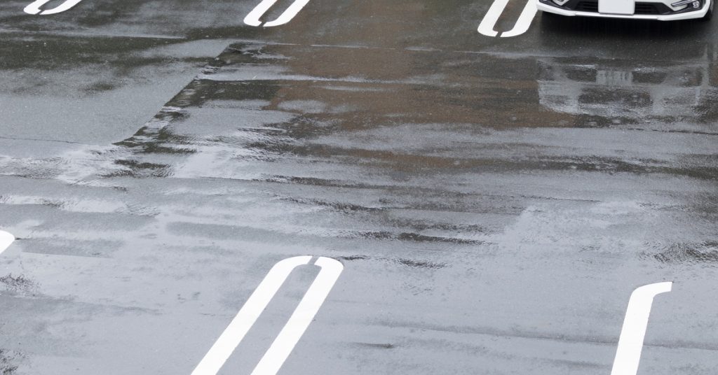 A wet parking lot after a rainstorm with one white car parked in one of its spaces.