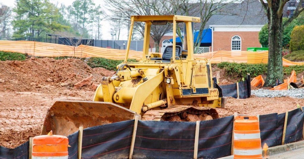 An unattended yellow bulldozer sits at a construction site behind a temporary fence and orange traffic barrels.