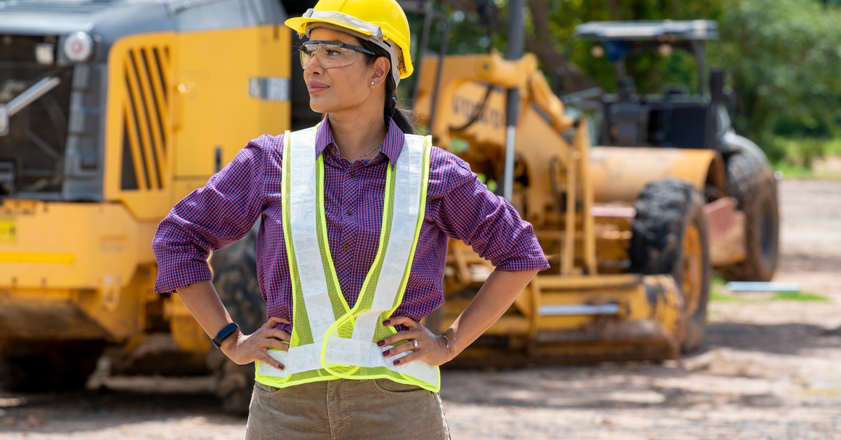 A woman in a yellow vest and hard hat stands in front of a bulldozer on the dirt ground of a construction site.