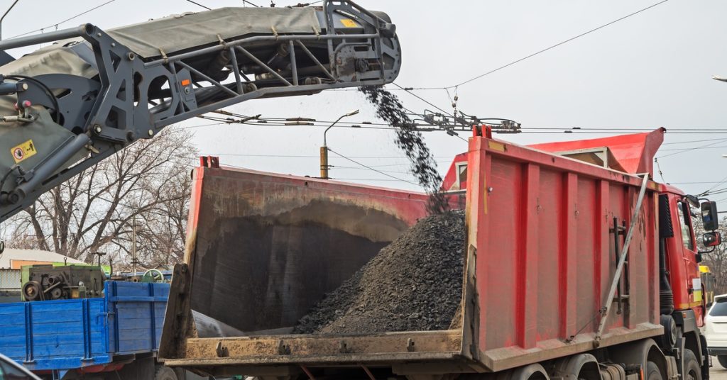 A large red dump truck is loaded with crushed asphalt from a milling machine during a road paving project.