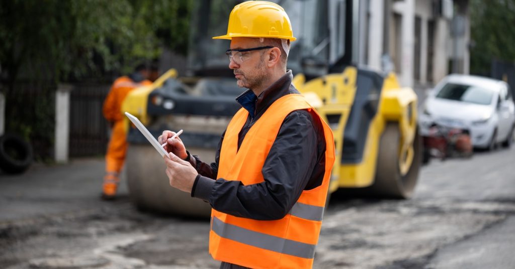 A man in an orange safety vest and hard hat writes on a clipboard while standing near a road undergoing maintenance.