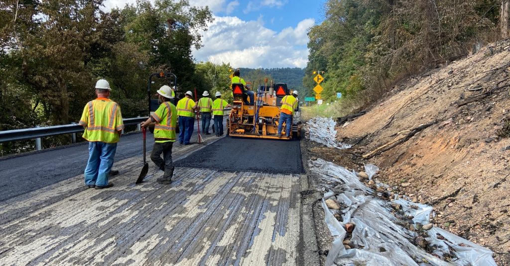 A construction crew in yellow safety vests stands on a road that's being repaved by an orange machine.
