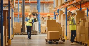 A warehouse worker waves to a coworker while pushing a forklift with cardboard boxes. Another worker is in front of them scanning boxes.
