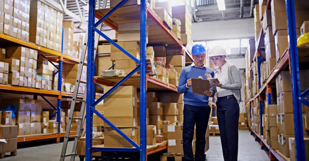 A man and a woman in professional attire and hard hats standing inside a warehouse and looking at a clipboard together.