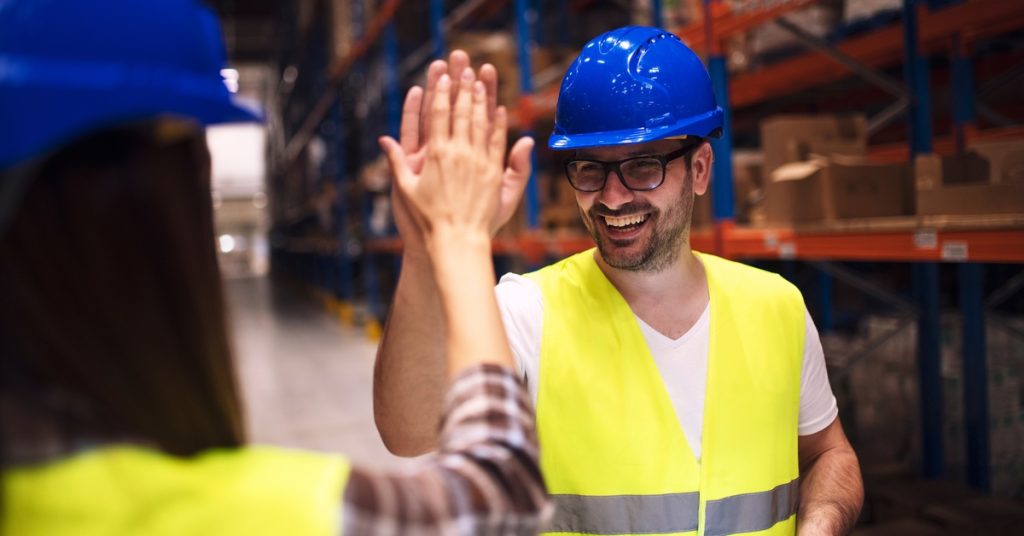 Two warehouse workers in yellow safety vests and blue hard hats giving each other a high five while standing between two shelves in a warehouse.
