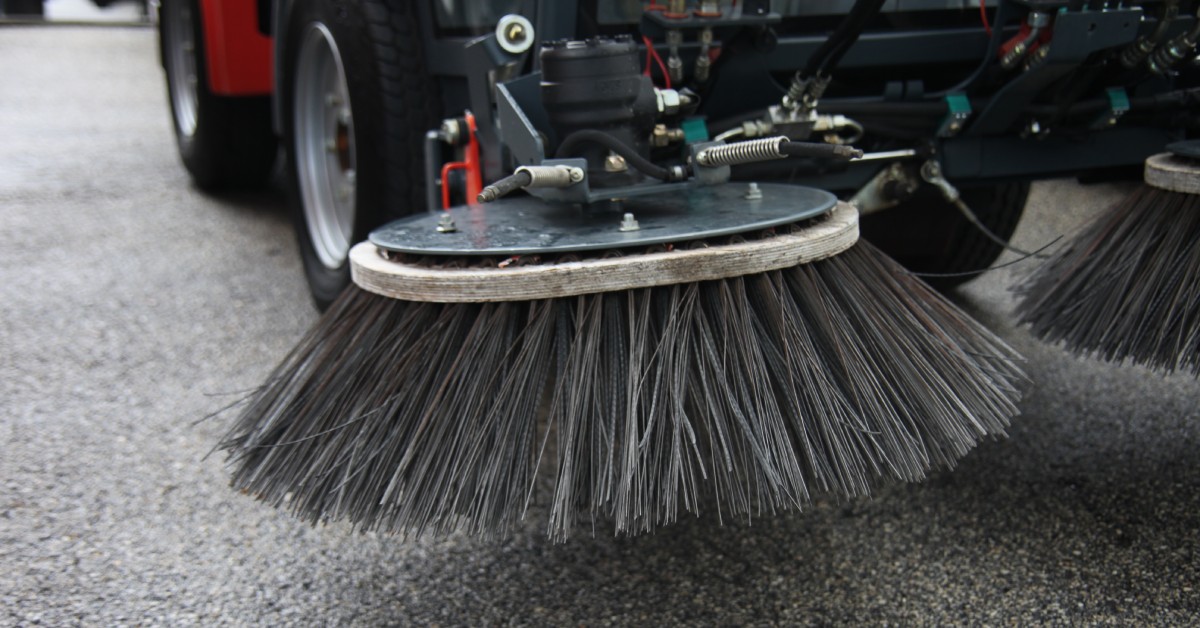 A parked street sweeping vehicle has round brushes with black bristles hovering above the clean pavement.
