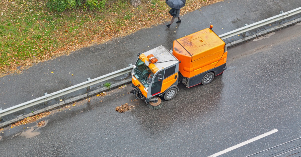 An orange street sweeping truck drives across a road with its brushes cleaning off the asphalt surface.