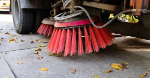 A commercial street sweeping vehicle has circular brushes with red bristles held above a street with leaves.