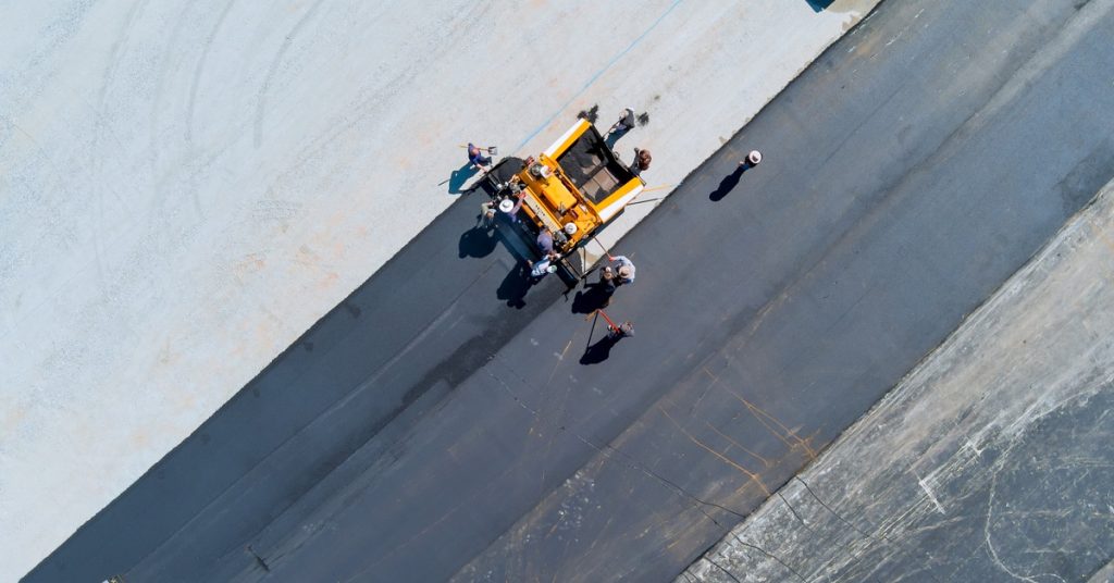 A group of road repair workers stand around a yellow machine as it repairs a large parking lot, turning pavement black.