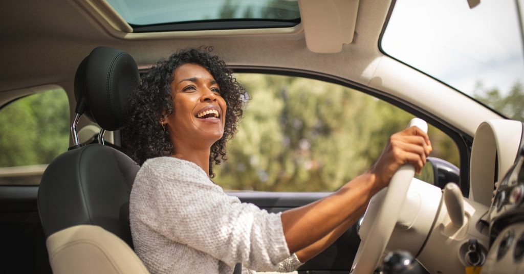A woman smiles and looks up toward her right side while driving a car. She has both her hands on the wheel.