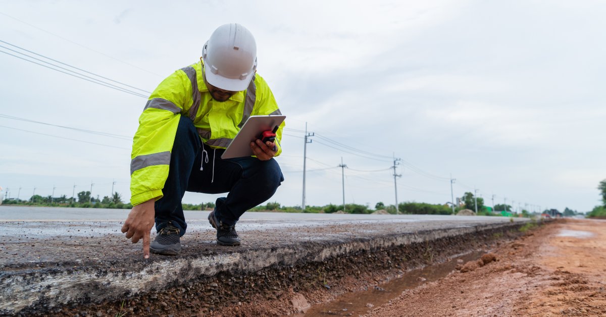 A construction worker wearing a yellow vest and a hard hat squats down to touch the edge of the asphalt road.