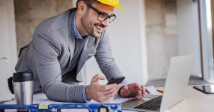 A smiling man in professional attire and a yellow hard hat types on a laptop and holds a smartphone.