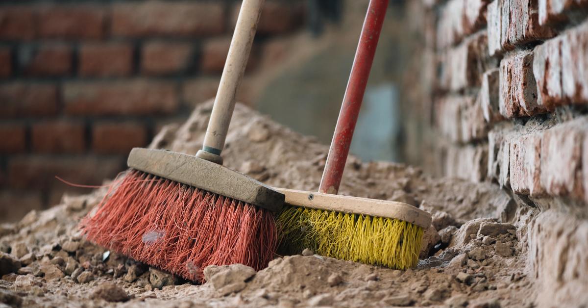 A broom with red bristles and one with yellow bristles lead against a brick wall on top of a large dirt pile.
