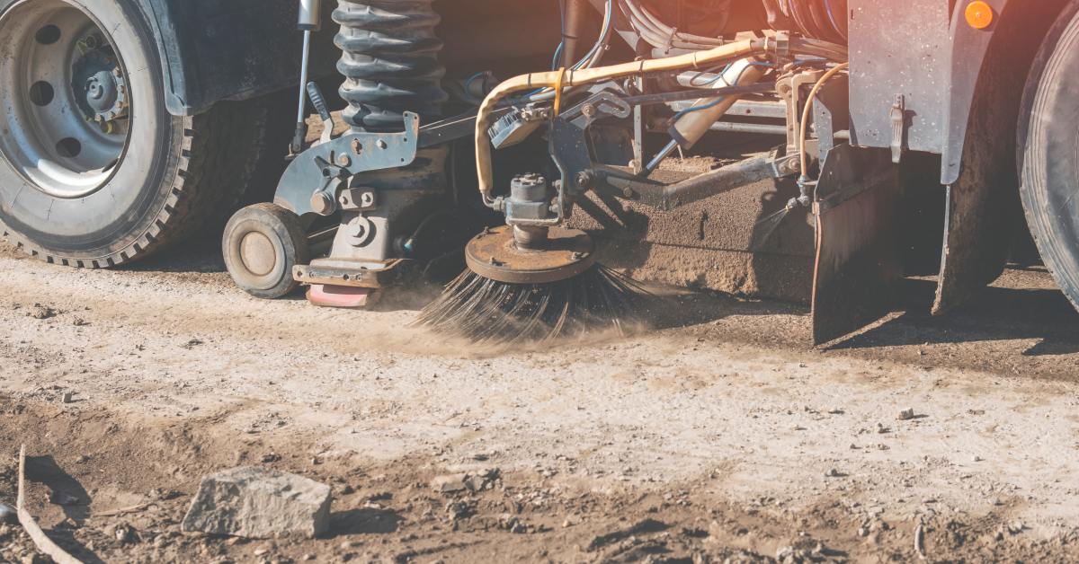 The circular black bristle brush of a street-sweeping machine rotates on top of the dirt road of a construction site.