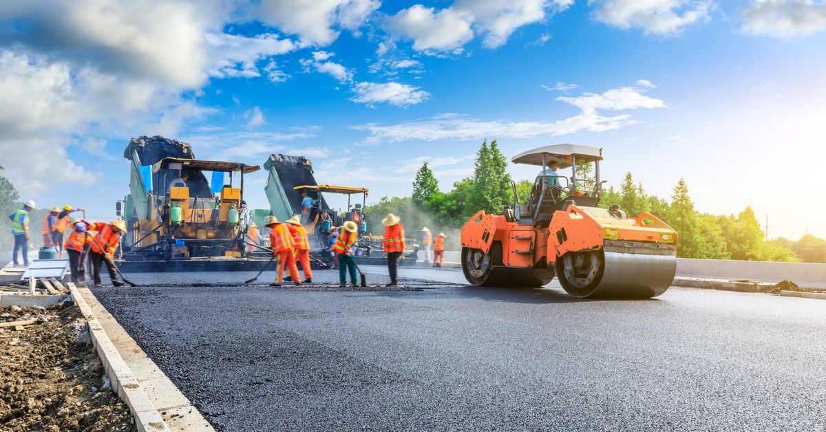 A construction crew, all members dressed in orange outfits and standing with heavy machinery, work on the street.