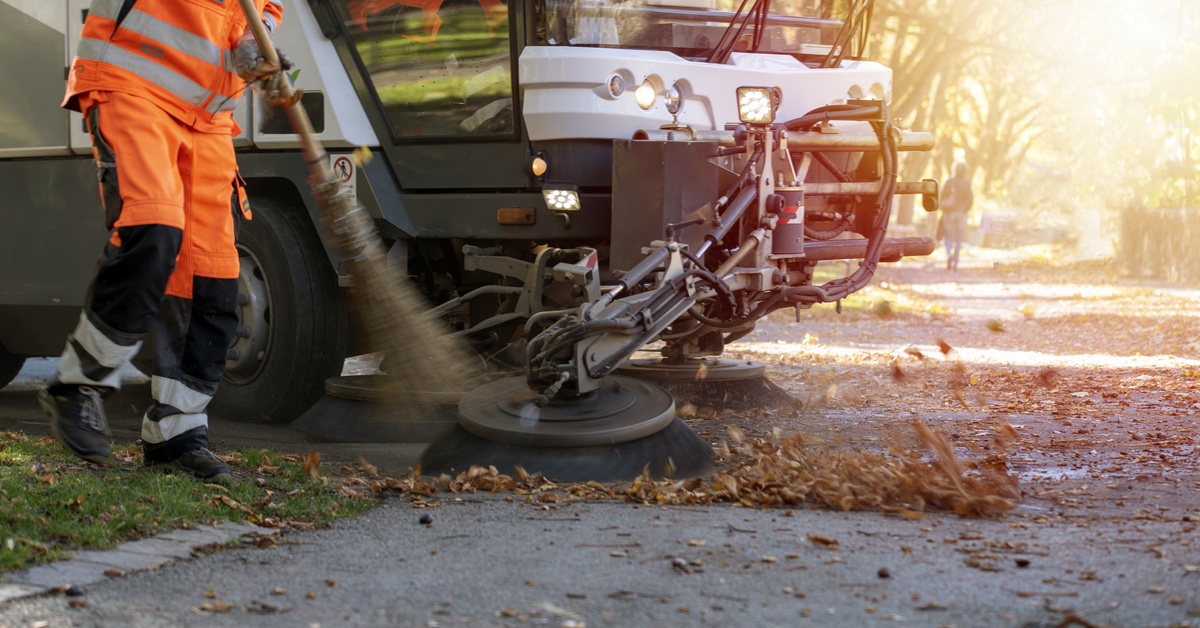 A man in an orange uniform sweeps the leaves on the street with a broom next to a street sweeping machine.
