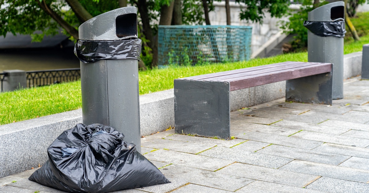 A filled black trash bag sits on the ground next to a public trash can next to a bench on the side of the street.