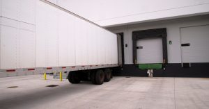 A large white truck trailer sits parked in a warehouse loading dock next to a couple of unused doors.