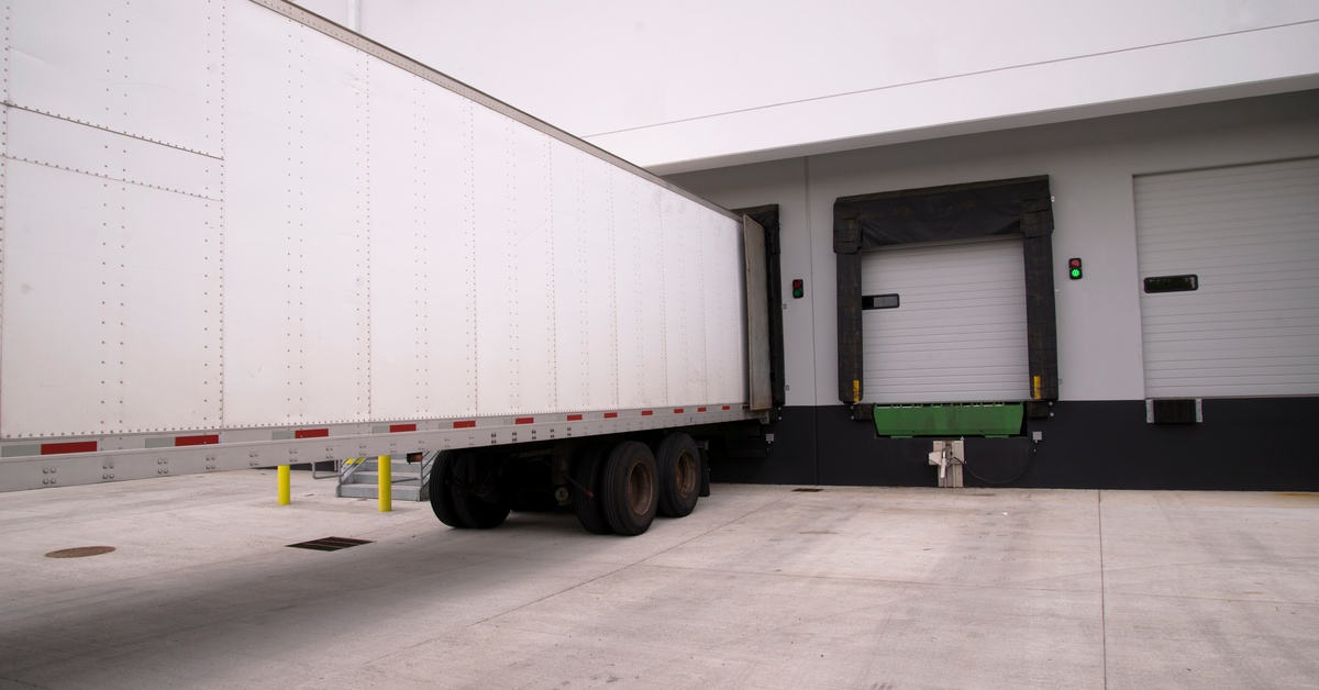 A large white truck trailer sits parked in a warehouse loading dock next to a couple of unused doors.