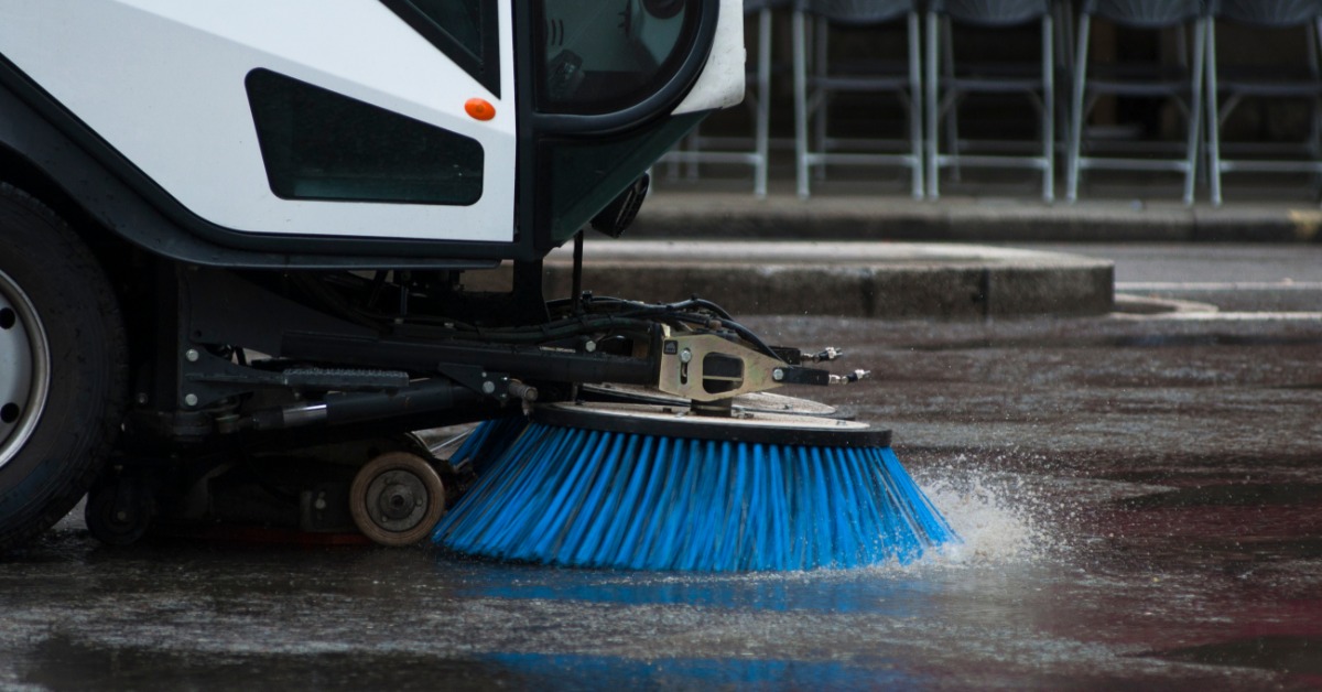 A white street sweeping vehicle has a large, circular brush with blue bristles. There are fold-out chairs in the background.